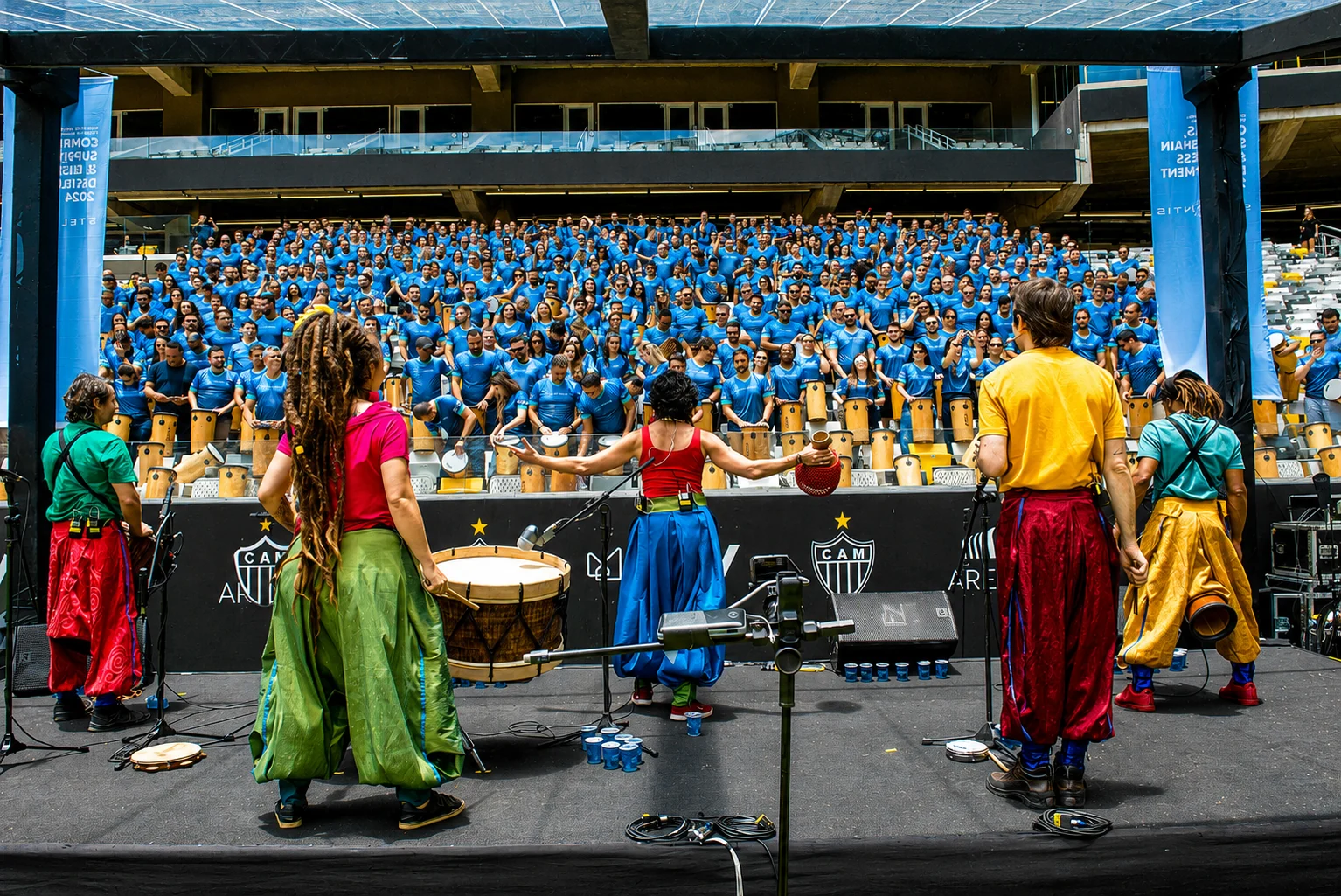 Os artistas do Grupo Trampulim de frente para uma arquibancada lotada, cada pessoa com um tambor, na Arena MRV